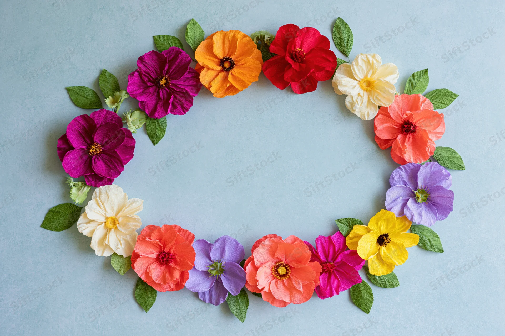 Handmade crepe paper flowers arranged in an oval wreath frame on a light blue background