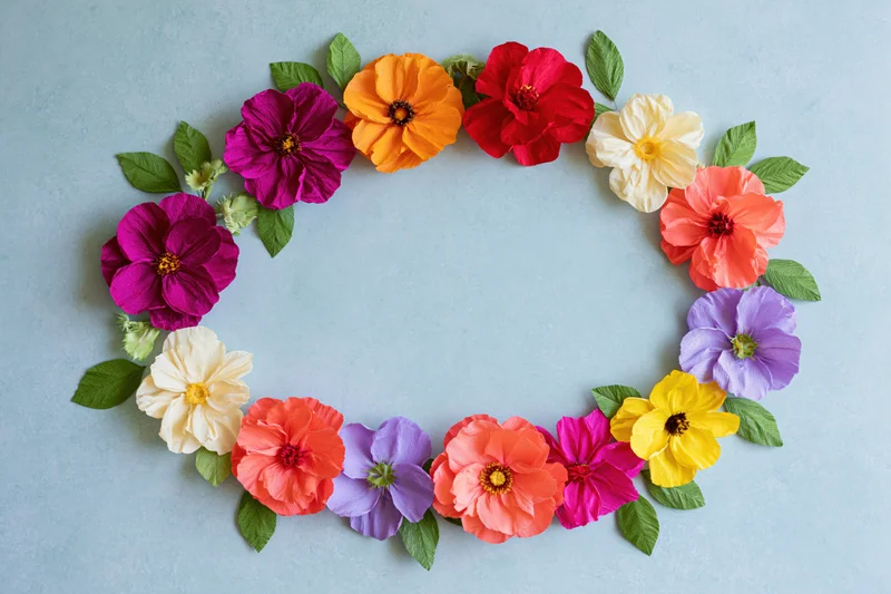 Handmade crepe paper flowers arranged in an oval wreath frame on a light blue background