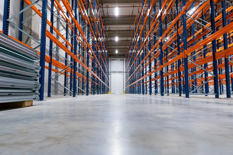 Empty warehouse interior with tall blue and orange metal shelving units lining a concrete aisle