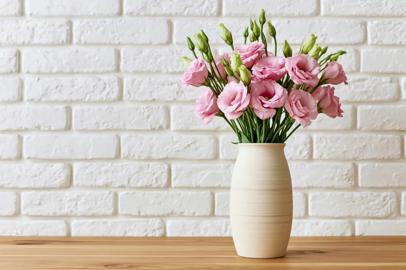 Matte beige ceramic vase holding a loose bouquet of pink lisi flowers on a wooden table against a white brick wall