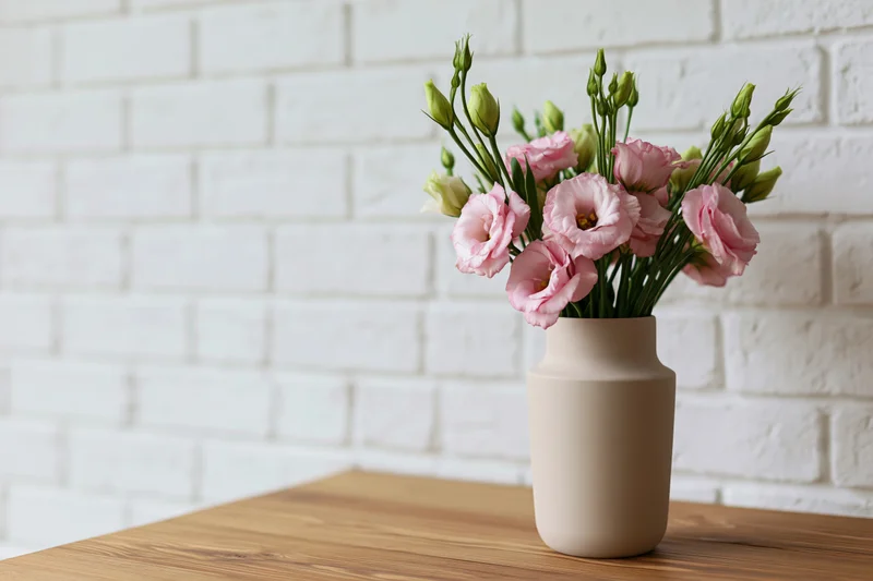 Matte beige ceramic vase holding a loose bouquet of pink lisianthus flowers on a wooden table against a white brick wall