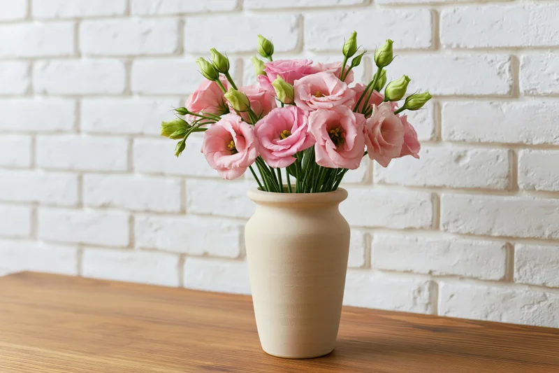 Matte beige ceramic vase holding a loose bouquet of pink lisianthus flowers on a wooden table against a white brick wall