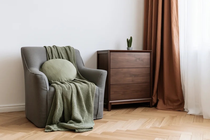 Gray armchair with green blanket and cushion beside walnut dresser in bedroom corner near window with brown curtains