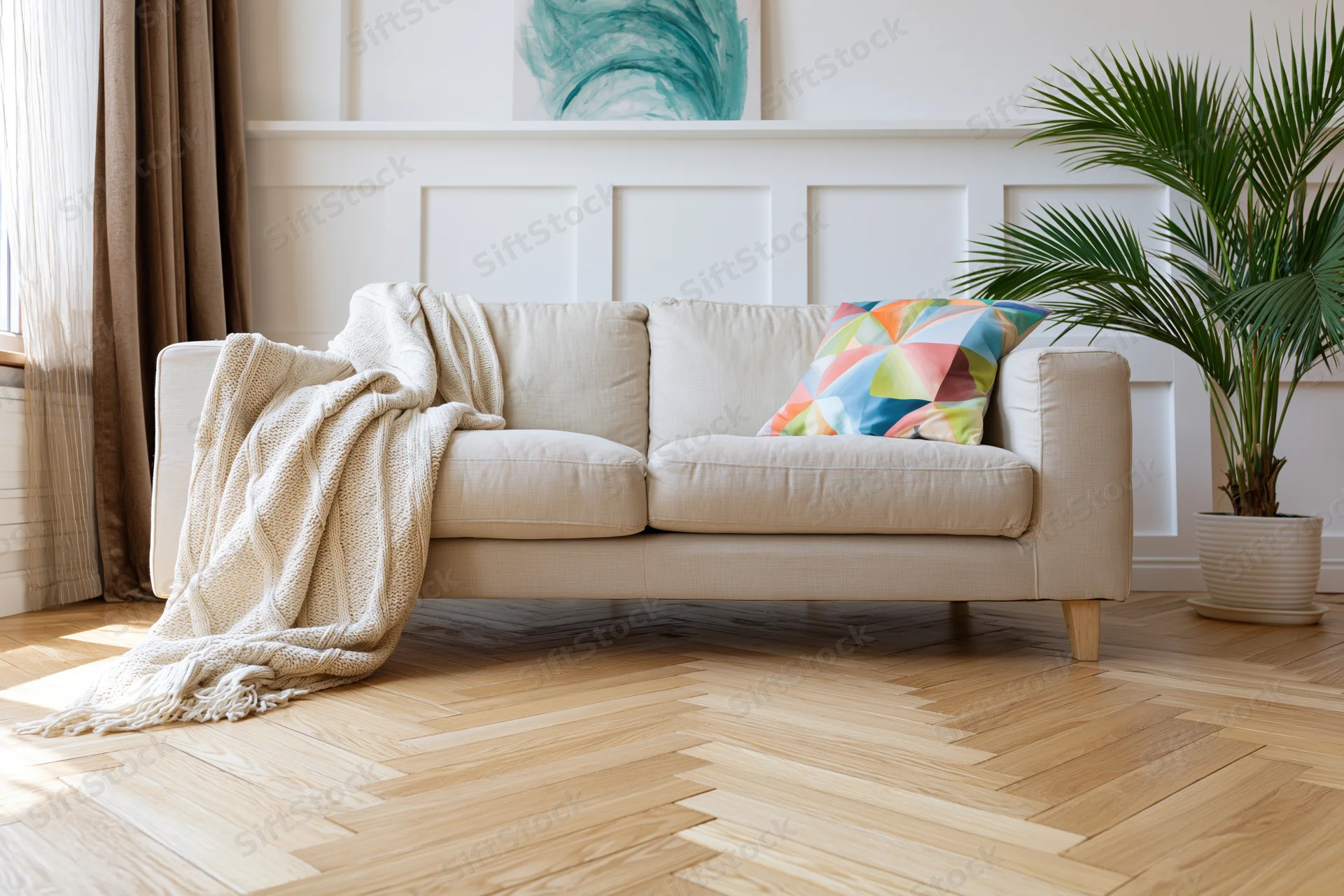 Beige linen sofa with colorful pillow and knitted throw in minimalist living room with wooden parquet floor and potted palm plant