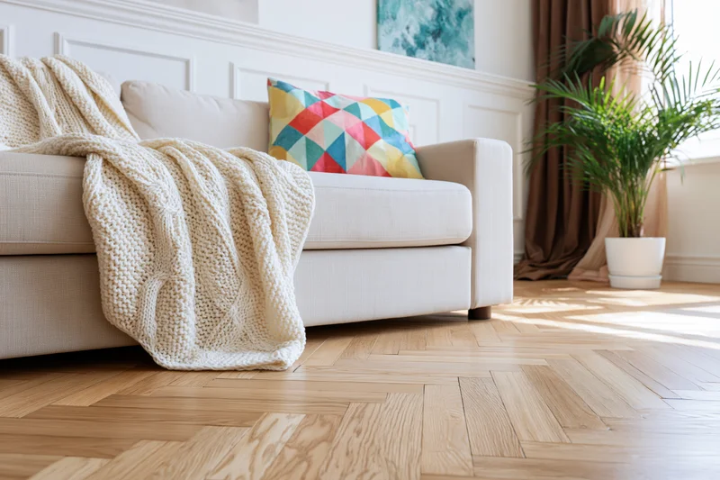 Beige linen sofa with colorful geometric pillow and knitted throw in minimalist living room with parquet flooring and potted palm near window