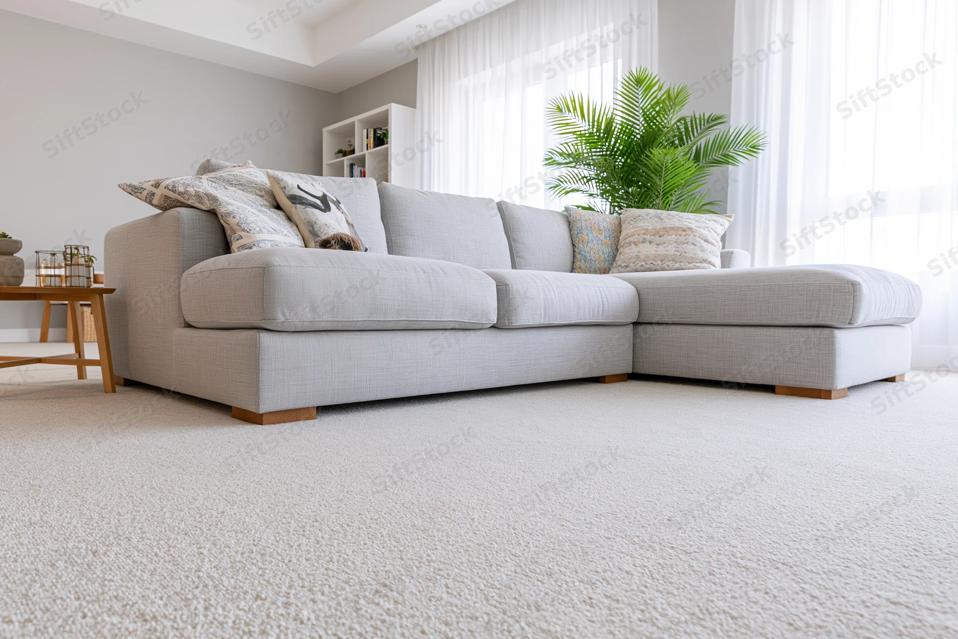 Low-angle view of a modern minimalist living room with a light gray sectional sofa, white carpet, and sheer curtains over a window, featuring decorative pillows and a potted palm plant