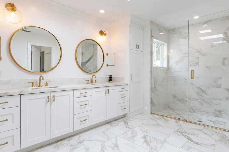 Modern primary bathroom with double vanity and round mirrors above marble countertop featuring gold fixtures and glass shower enclosure