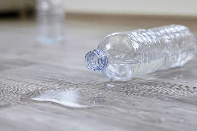 Overturned clear plastic water bottle lying on its side with spilled liquid on wooden floor