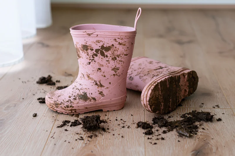 Muddy pink rubber rainboots left on light wood floor with scattered dirt