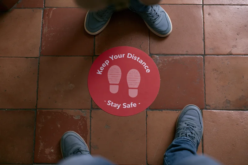 Two people standing on either side of a red circular floor sticker with footprints and text promoting social distancing in a tiled indoor space