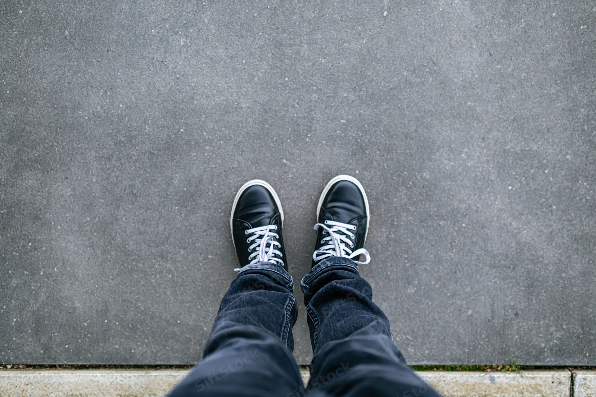 Person standing still on flat concrete pavement viewed from above, wearing black sneakers and jeans