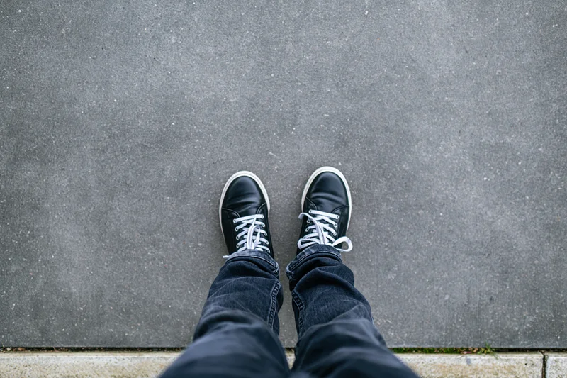 Person standing still on flat concrete pavement viewed from above, wearing black sneakers and jeans