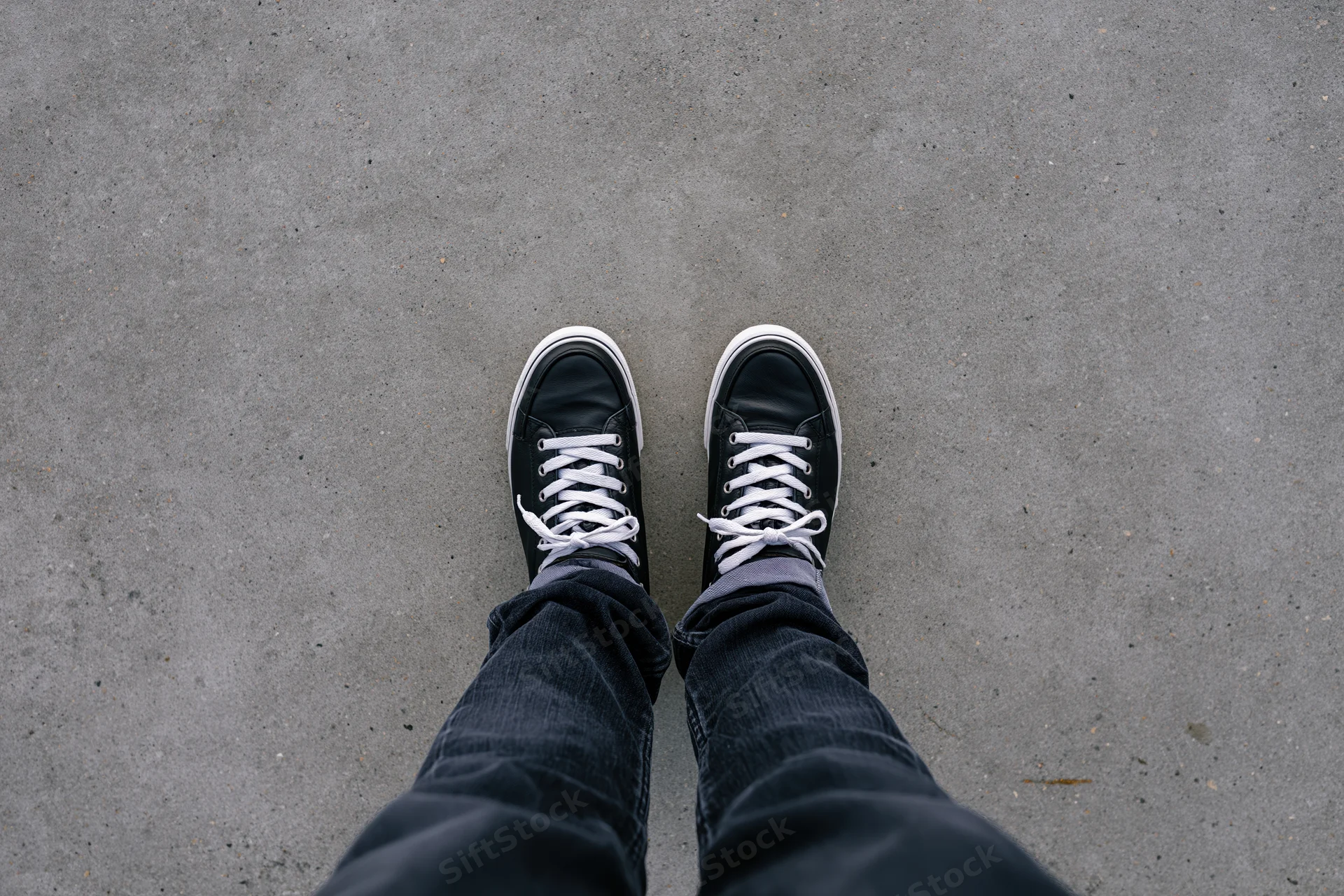 Person standing still on flat concrete pavement top-down view wearing black sneakers and jeans