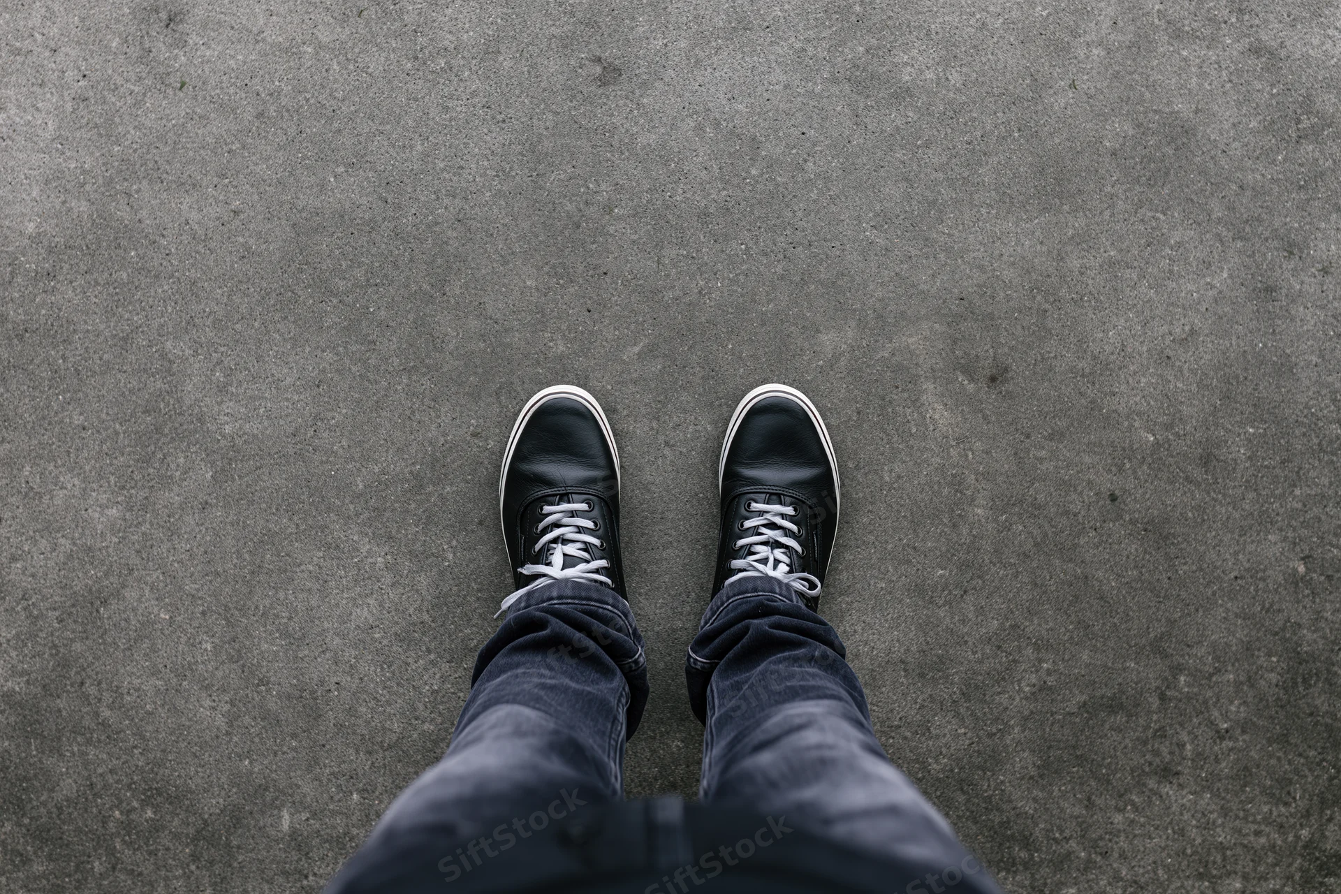 Person standing still on flat concrete pavement viewed from above, wearing black sneakers and jeans