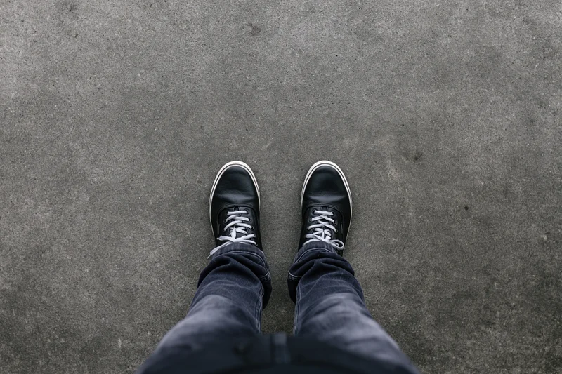 Person standing still on flat concrete pavement viewed from above, wearing black sneakers and jeans