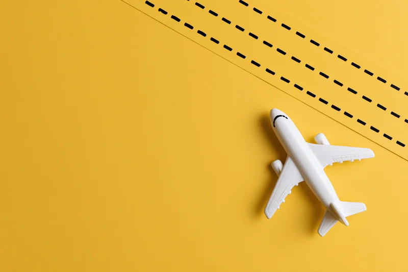 White plastic toy airplane lying flat on a solid yellow surface with a black dashed line running diagonally across the background