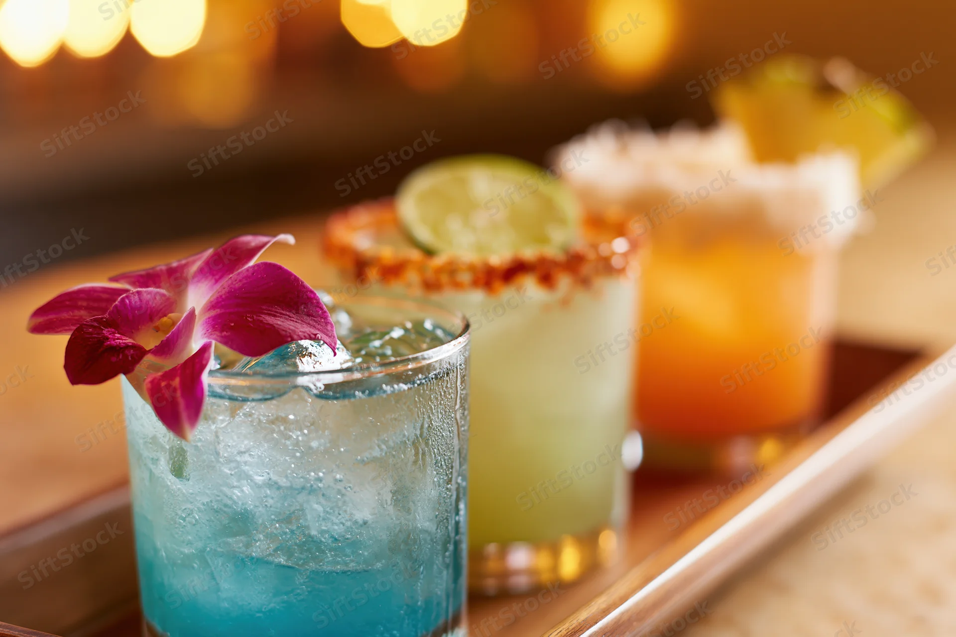 Three tropical cocktails lined up on a wooden bar tray with garnishes and ice
