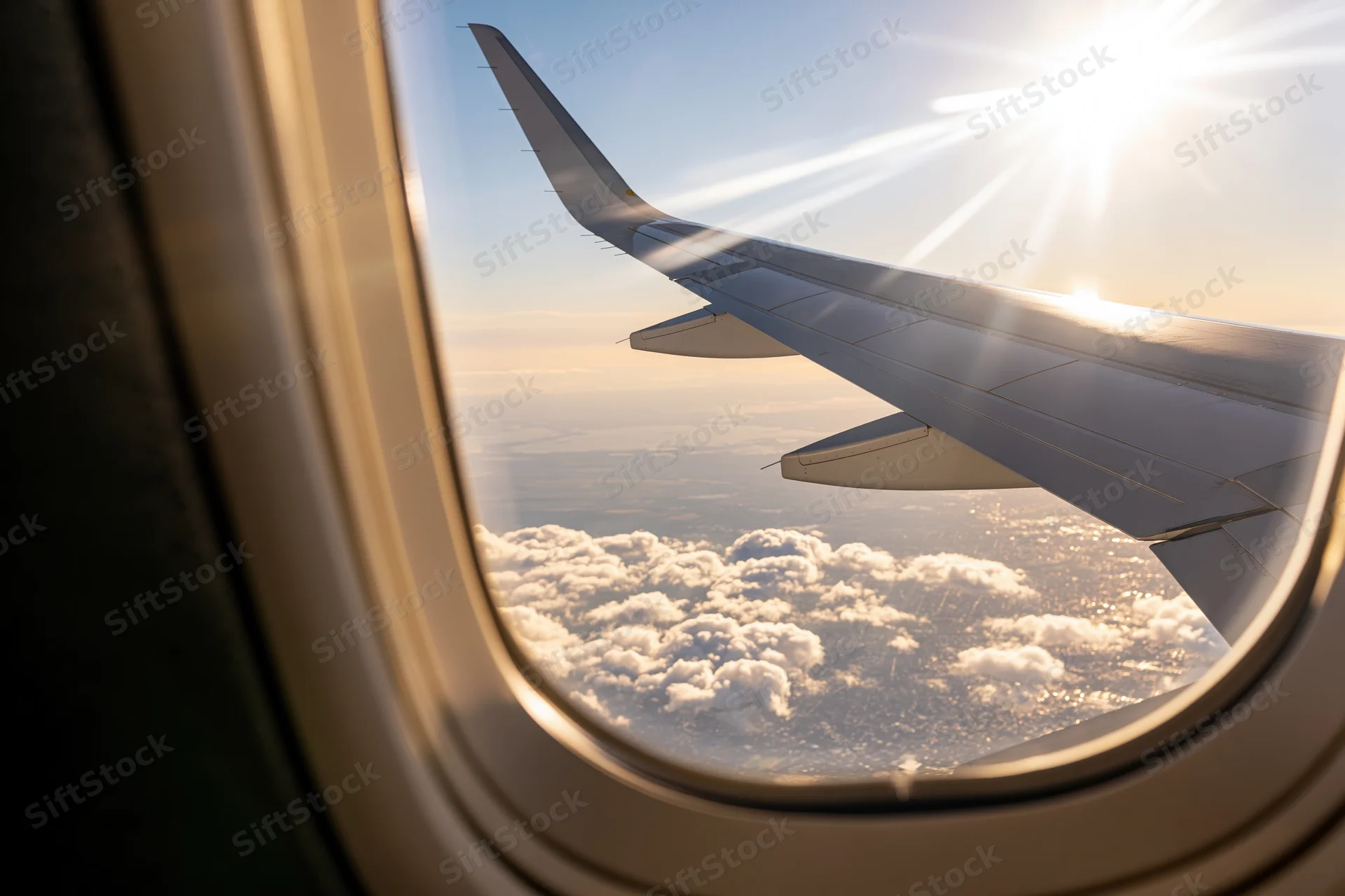 View through airplane window showing wing and clouds under bright sun at altitude