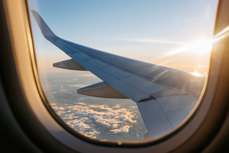Airplane wing seen through oval window during sunset flight above clouds