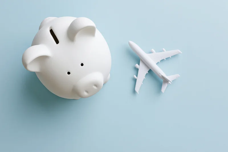 White ceramic piggy bank next to a white airplane model on light blue surface
