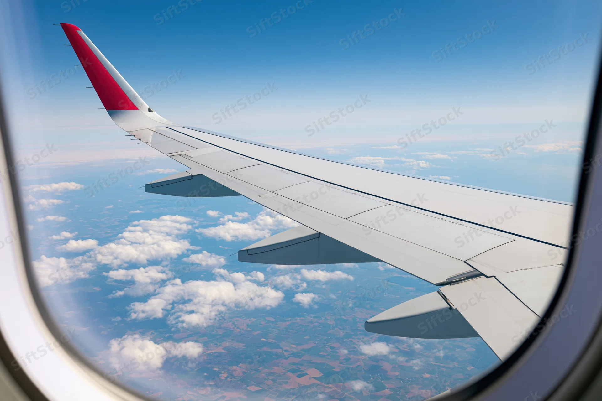 Airplane wing with red wingtip seen through window above clouds and rural landscape