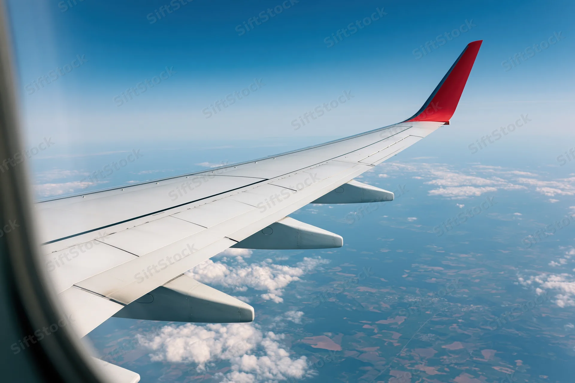 Airplane wing with red wingtip viewed from window during flight over clouds and landscape