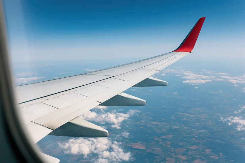 Airplane wing with red wingtip viewed from window during flight over clouds and landscape