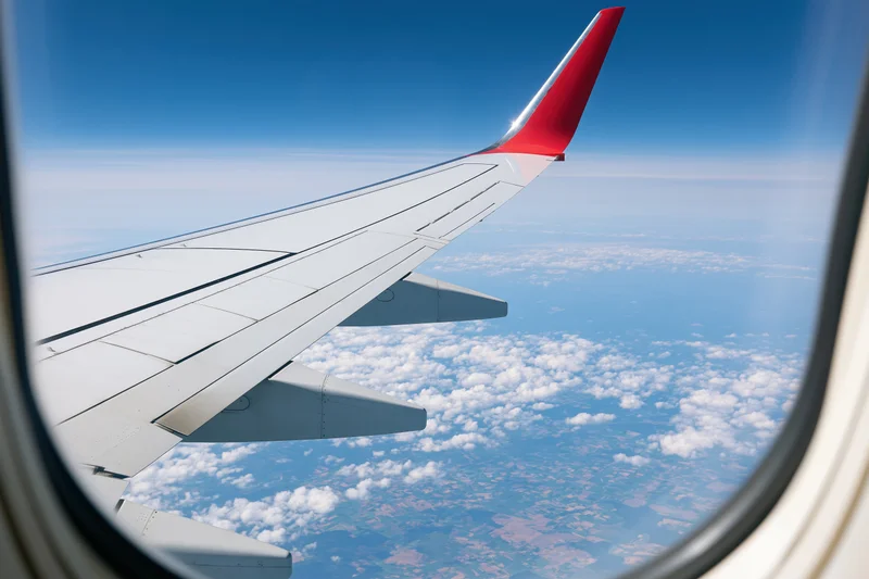 White commercial airplane wing with red wingtip seen through window above clouds