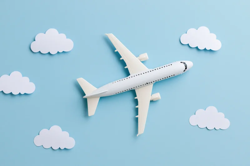White toy passenger airplane model flying among paper clouds on light blue background