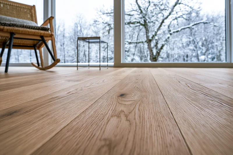 Wide-plank oak hardwood floor filling the foreground with a snowy winter landscape visible through large windows in a modern living room interior