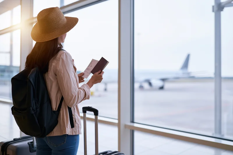 Young woman holding passport and ticket at airport terminal window with luggage and airplane view