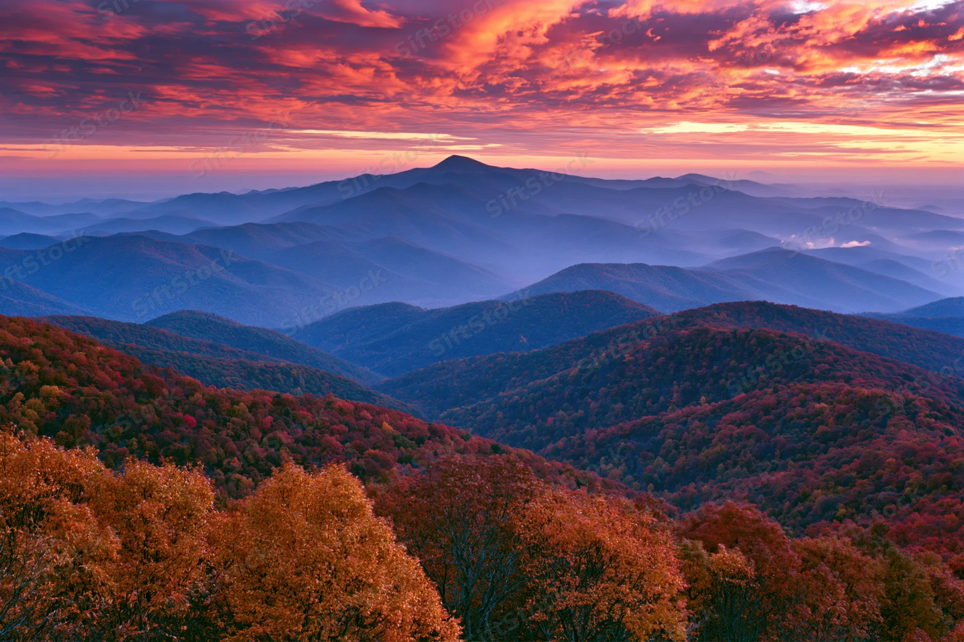 Rolling mountains covered in autumn foliage under a fiery sunset sky
