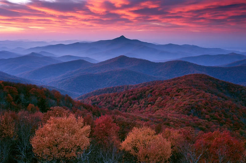 Rolling mountains covered in autumn foliage under a colorful sunset sky