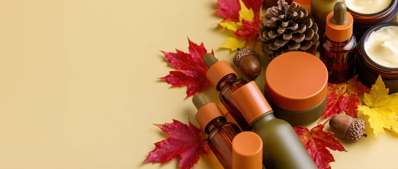 Autumn skincare products arranged with leaves and pinecones along right edge on beige background