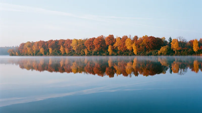 Autumn trees in peak fall color lining the far bank of a calm lake with their reflection mirrored in the still water