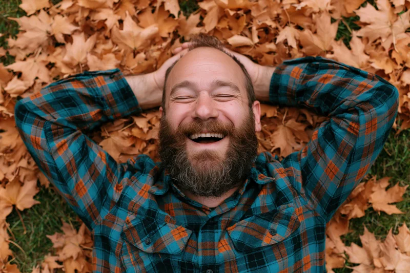 Bearded man laughing with hands behind head lying on autumn leaves in a grassy outdoor area