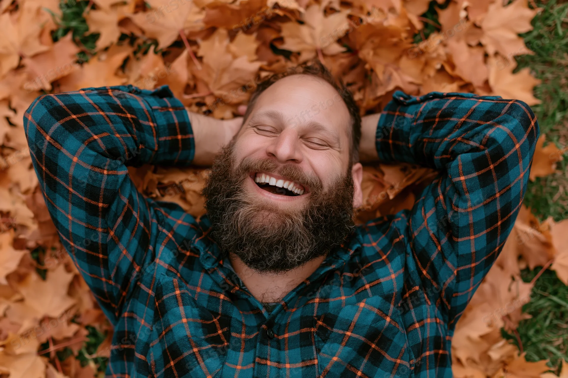 Bearded man laughing with eyes closed lying on back in pile of autumn leaves