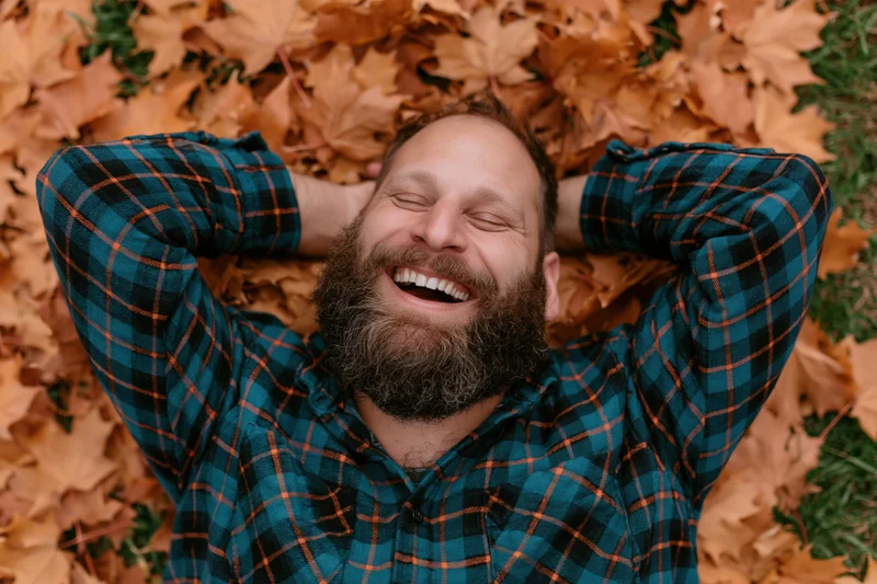 Bearded man laughing with eyes closed lying on back in pile of autumn leaves