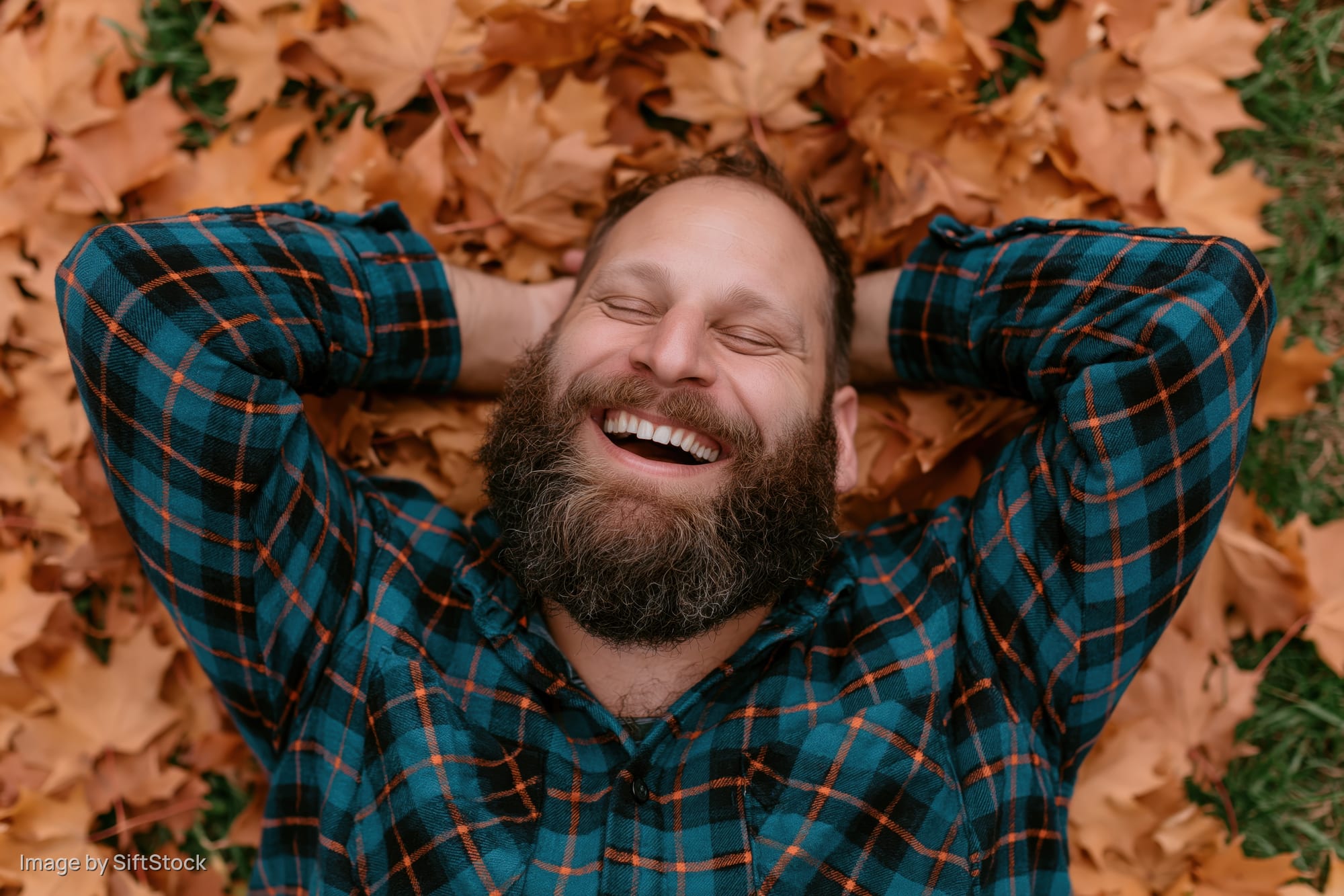 Bearded man laughing with eyes closed lying on back in pile of autumn leaves