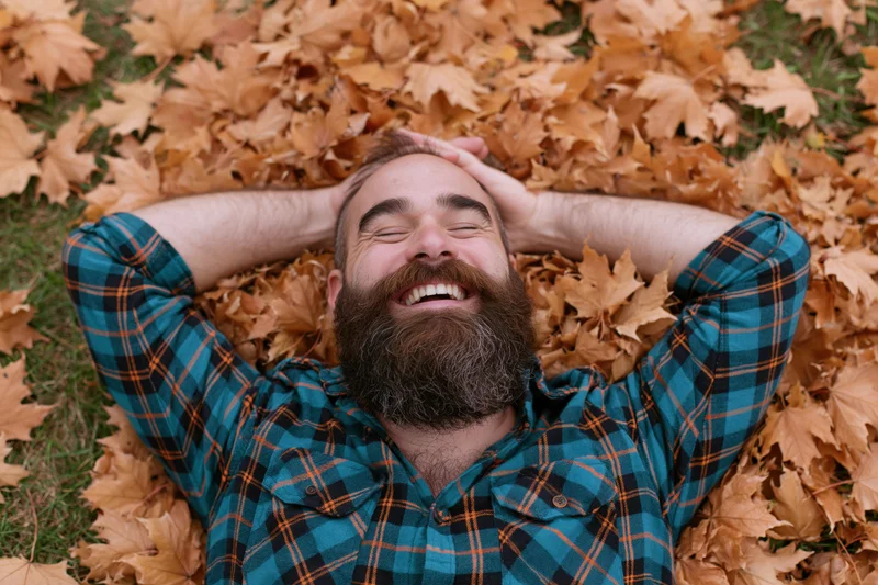Bearded man laughing while lying on his back in a pile of autumn leaves outdoors