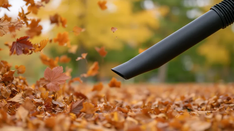 Black leaf blower nozzle blowing autumn leaves across a yard covered in fallen foliage