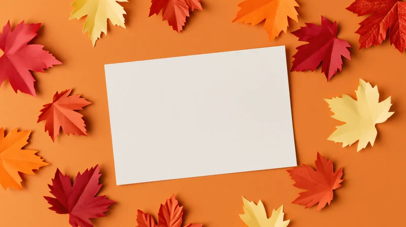 Blank white card centered on orange surface surrounded by colorful autumn maple leaves in flat lay arrangement