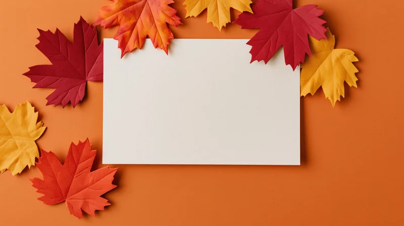 Blank white card surrounded by colorful autumn maple leaves on orange background
