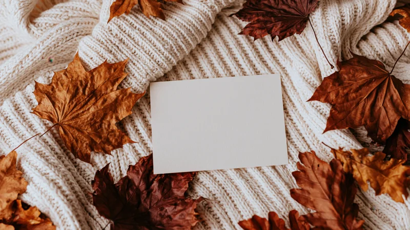 Blank cream card lying on cream ribbed knit sweater with scattered autumn maple leaves