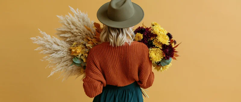 Woman from behind holding large autumn bouquet of dried pampas grass and colorful flowers against warm yellow backdrop
