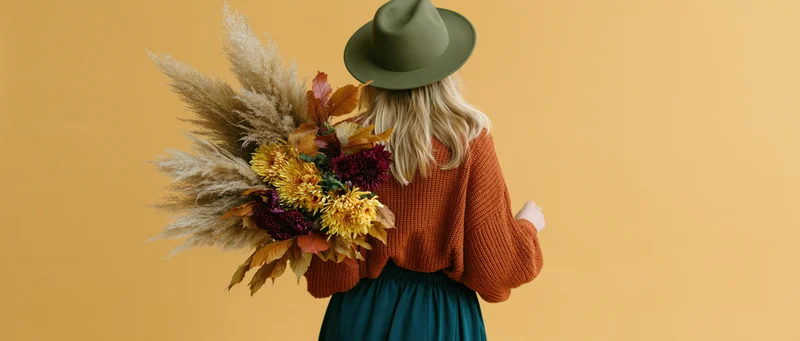 Woman from behind holding large autumn bouquet with pampas grass and colorful flowers against warm yellow background