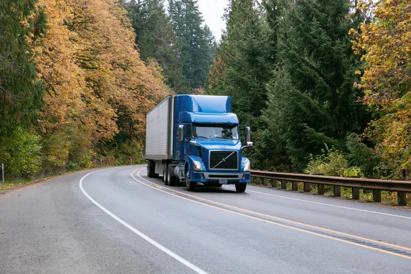 Blue semi-truck with white refrigerated trailer driving along curved highway through autumn forest