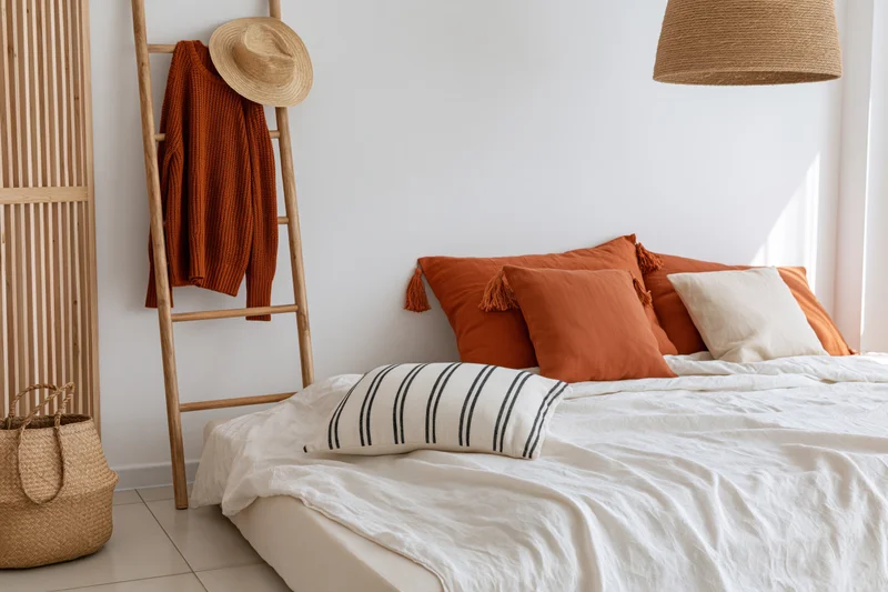 Low platform bed with white bedding and orange pillows in a bohemian bedroom with wooden ladder, woven hat, and wicker basket