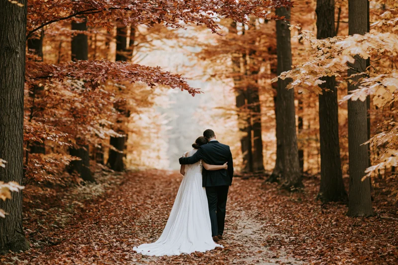 Bride and groom embracing from behind on a leaf-covered forest path in autumn