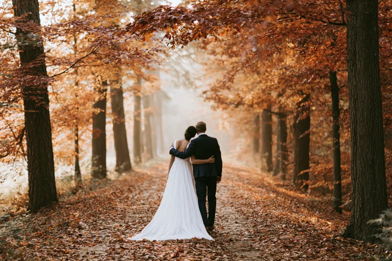 Bride and groom walking away together down a leaf-covered forest path in autumn